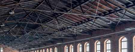 Foto de Gable roof truss of a large, vintage factory hall. Roofing construction (sheathing) made of wooden planks. Brick walls and arcade windows. Industrial interior. - Imagen libre de derechos