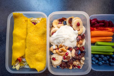 Boxes with healthy food full of colorful vegetables and fruits. Nuts, blueberries, yogurt, pomegranates, bananas, gluten-free pancakes, raisins, celery, carrots and peppers. Meals for the day. 