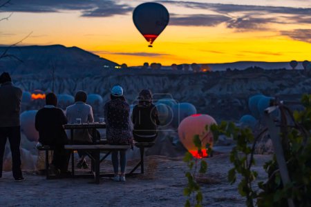 Téléchargez les photos : 10.11.2022 Cappadoce, Turquie. Groupe de divers touristes assis sur un banc sur une colline et regardant décoller étonnamment des montgolfières en Cappadoce, Turquie. Beau lever de soleil, ciel orange. Haut - en image libre de droit