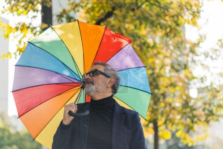 Téléchargez les photos : Homme d'âge moyen aux cheveux gris et barbe portant des lunettes et un blazer tenant un parapluie arc-en-ciel regardant autour du parc souriant. Plan horizontal extérieur. Photo de haute qualité - en image libre de droit