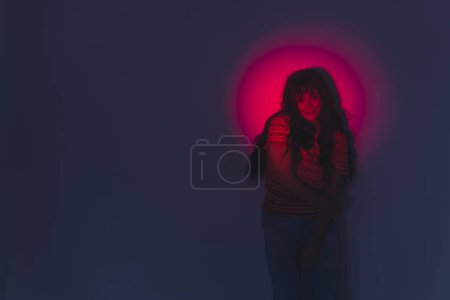 Téléchargez les photos : Photoshoot lampe virale esthétique moderne. Projecteur de lumière rouge illuminant le visage et les bras de la belle jeune fille adulte en t-shirt rayé. Cheveux longs, noirs, ondulés avec frange. Prise de vue. Haute qualité - en image libre de droit