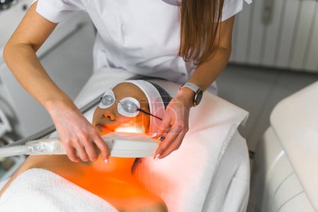 Beautiful Asian young adult woman during thermolifting treatment at her favorite SPA performed by unrecognizable caucasian beautician. Protective gear and spa interior. High quality photo
