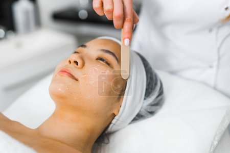 Téléchargez les photos : Belle femme asiatique allongée sur un lit SPA pendant la procédure de soins apaisants. Esthéticienne professionnelle méconnaissable utilisant un bâton de bois pour appliquer un masque transparent ou un produit nettoyant. Gros plan - en image libre de droit