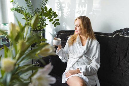 Photo for Wealthy caucasian blond woman in soft white robe sitting on exclusive black sofa among flowers and drinking coffee or tea from a white cup. SPA day and relaxation. High quality photo - Royalty Free Image