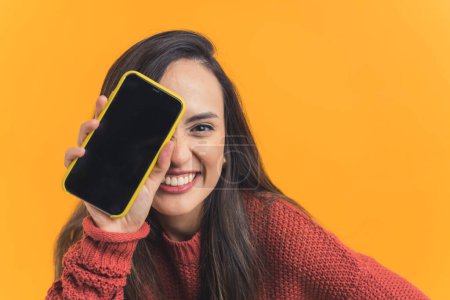 Nett aussehendes lateinamerikanisches, braunhaariges Mädchen, das seinen Handybildschirm in die Kamera zeigt, mittlere Nahaufnahme isoliert orangefarbenen Hintergrund. Hochwertiges Foto