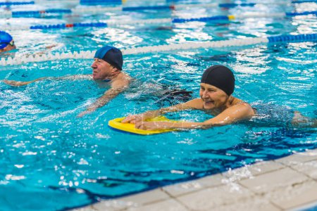 Indoor activities for well-being. Grandmother and grandfather swimming next to each other and wearing protective head caps. Sports pool interior and equipment. High quality photo