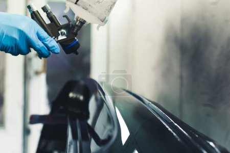 Car renovation and detailing process. Close-up perspective of car part being varnishing in paint spray booth by a hand in a protective glove holding spray gun. High quality photo