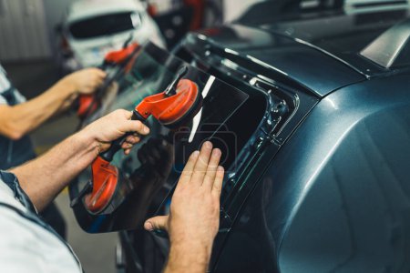 Automobile repair process. Closeup indoor portrait of two unrecognizable caucasian blue collar workers using suction cups to remove rear window from a black car. High quality photo