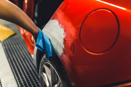 Hand of unrecognizable caucasian mechanic person using blue cloth to smooth out the surface of red car covered with automotive paint before varnishing . High quality photo