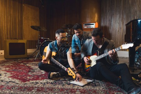 Foto de Dos jóvenes guitarristas y un baterista sentados en el suelo y mirando la hoja de notas sobre la repetición, el concepto de música de estudio de grabación. Foto de alta calidad - Imagen libre de derechos