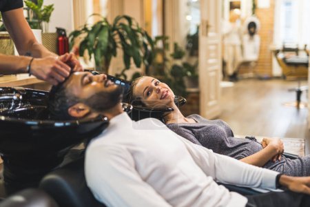 Téléchargez les photos : Client indien se faisant laver les cheveux par un coiffeur à côté d'une cliente blonde heureuse dans un salon de coiffure professionnel. Photo de haute qualité - en image libre de droit