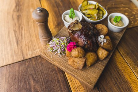 Photo for Meat dish in traditional Polish cuisine. Golonka z pieca - roasted pork knuckle with roasted potatoes, sauces, pickles and pepper shaker on wooden table. Top view. High quality photo - Royalty Free Image