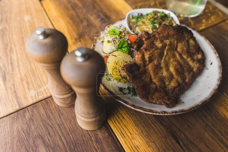 Photo for Polish restaurant dish schabowy - pork chop with potatoes and vegetables served on a white plate along with pepper and salt shaker. High angle view. Indoor shot. High quality photo - Royalty Free Image
