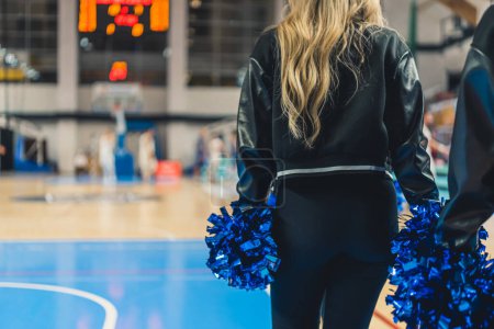 Foto de A cheerleader from the back in a stadium during a match. She is holding pom poms in her hands. The girl is dressed in a black jacket and fitted trousers. Players can be seen in the background. - Imagen libre de derechos