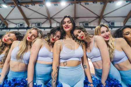 Téléchargez les photos : Group of cheerleaders is captured in a playful pose, resting their heads on fellows shoulders as they smile for the camera. Cheerleaders are dressed in blue uniforms and each holds a pair of pom-poms - en image libre de droit