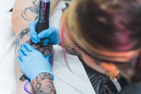 Focused female tattoo artist doing her freehand tattoo on her clients leg. Blurred artist head in the foreground. Holding professional tattooing equipment. High quality photo