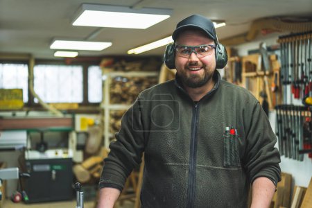Photo for Happy male carpenter with protective glasses and headphones on, workshop background. High quality photo - Royalty Free Image