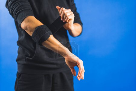 Téléchargez les photos : Une personne enveloppant un support de coude pour réduire la douleur, fond bleu main blessée. Photo de haute qualité - en image libre de droit