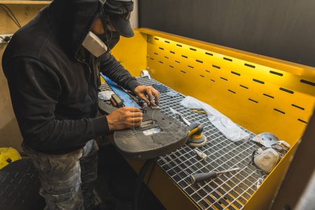 Mann mit Schutzmaske poliert Kohlefaser-E-Gitarrenkörper in der Herstellung. Hochwertiges Foto