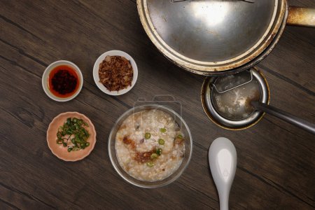 Asian thick rice porridge bubur chicken beef meat fish prawn rustic silver pot ladle serving in glass bowl fried shallots green onion chilli oil condiment 