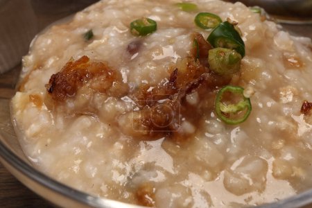 Asian thick rice porridge bubur chicken beef meat fish prawn rustic silver pot ladle serving in glass bowl fried shallots green onion chilli oil condiment 