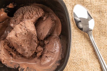 Photo for Dark brown chocolate ice cream in a black ceramic bowl with a metal spoon on a jute napkin, close-up, top view. - Royalty Free Image