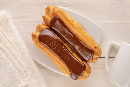 Two chocolate eclairs with white ceramic saucer and cup on wooden table, close-up, top view.