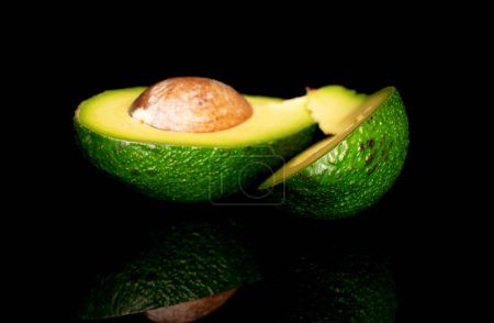 Photo for In the foreground, in focus, is one whole bone of a mature avocado. Behind it, in the background are two halves, cut along and lying on top of each other, a green , avocado, on a black background. - Royalty Free Image