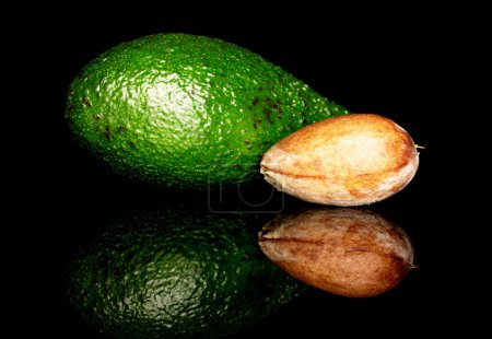 Photo for In the foreground, one round whole brown avocado pit is in focus. Behind it is one green ripe avocado. The background is black. - Royalty Free Image