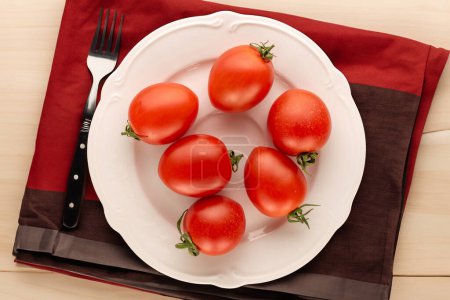 Foto de Varios tomates cóctel en un plato de cerámica blanca con una servilleta y un tenedor en una mesa de madera, macro, vista superior. - Imagen libre de derechos