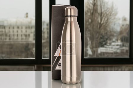 Photo for One metal thermos bottle with a paper box is on the table, close-up, against the background of the window. - Royalty Free Image