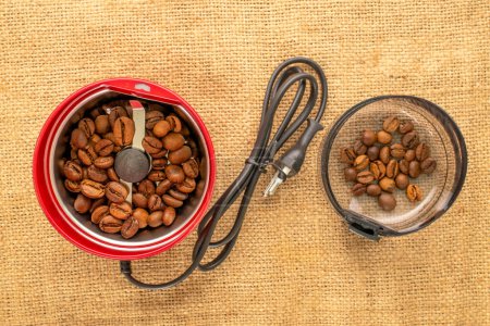 One electric coffee grinder with coffee beans on a jute cloth, macro, top view.