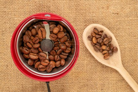 One electric coffee grinder with coffee beans and wooden spoon on jute cloth, macro, top view.