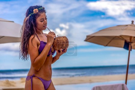 Foto de Mujer sexy disfrutando de un coco y festejando bajo los rayos del sol en una playa de arena blanca y dorada con el mar Caribe en el fondo, este es un lugar ideal para el turismo y las vacaciones de verano. - Imagen libre de derechos