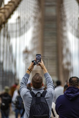 Photo for Man with mobile phone photographing the Brooklyn Bridge linking the boroughs of Manhattan and Brooklyn in New York City (USA), the largest suspension bridge in the world until 1889. - Royalty Free Image