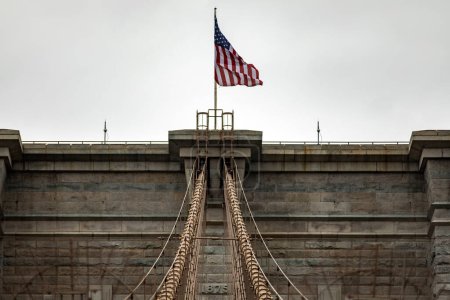 Photo for American flag on top of the Brooklyn Suspension Bridge linking the boroughs of Manhattan and Brooklyn in New York City (USA), the largest suspension bridge in the world until 1889. - Royalty Free Image