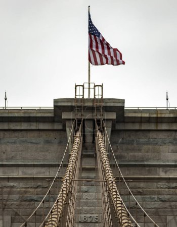 Photo for Photo of the American flag on top of the Brooklyn Suspension Bridge linking the boroughs of Manhattan and Brooklyn in New York City (USA), the largest suspension bridge in the world until 1889. - Royalty Free Image