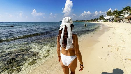 A sexy young woman enjoying a white sandy beach in the Riviera Maya, as she strolls along the pier over the crystal clear turquoise Caribbean water and heads to her resort in Mexico.