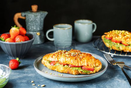 Photo for Croissant sandwiches or brioche filled with pistachio cream, strawberries, topped with caramel dip on plate and other backside. Grey background, ingredients, cups of coffee. Delicious breakfast - Royalty Free Image
