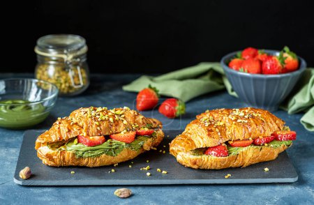 Photo for Two sweet croissant sandwiches or brioches filled with pistachio cream, strawberries, topped with caramel dip. Grey background, black stone board, green napkin, ingredients. Delicious breakfast - Royalty Free Image