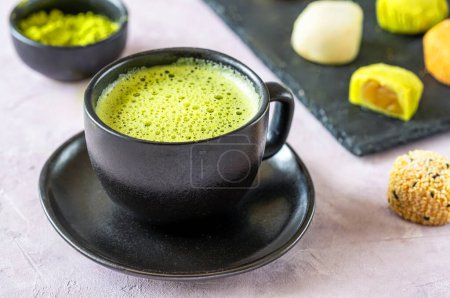 Una atractiva taza de té matcha espumoso servido en una elegante taza negra, con una selección de mochi y polvo de matcha suavemente borrosa en el fondo.
