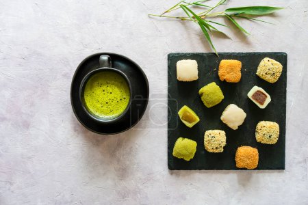 A serene arrangement of various mochi on slate with a cup of frothy matcha tea, set against a pale background with bamboo leaves
