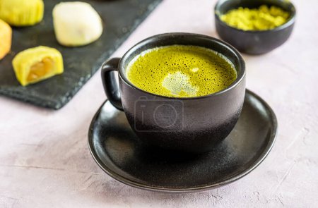 Nahaufnahme eines schaumigen grünen Matcha-Tees in einer schwarzen Keramiktasse mit Mochi-Desserts und Matcha-Pulver im Hintergrund.