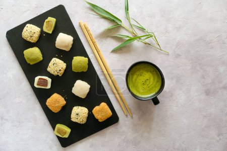 Assorted mochi on a dark slate board beside matcha tea and bamboo leaves, beautifully arranged on a textured backdrop