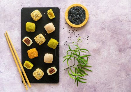 Bunte Auswahl japanischer Mochi auf schwarzem Schiefer mit Essstäbchen, schwarzem Sesam und Grün auf strukturiertem Hintergrund