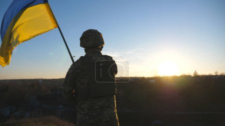 Soldier of ukrainian army stands at hill and holds waving flag of Ukraine. Man in military uniform and helmet lifted up flag against sunset. Victory against russian aggression. Invasion resistance.