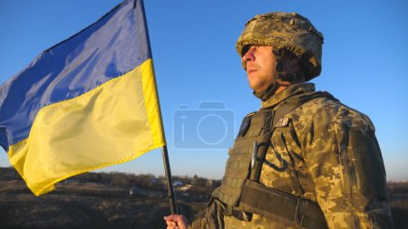 Photo for Ukrainian army soldier holding waving flag of Ukraine. Portrait of man in military uniform and helmet lifted up flag in hill. Victory against russian aggression. Invasion resistance concept. Slow mo. - Royalty Free Image