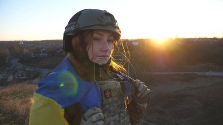 Female military medic of ukrainian army holding flag of Ukraine and looking into camera. Girl in uniform with yellow-blue flag happy by victory against russian aggression. Invasion resistance concept.