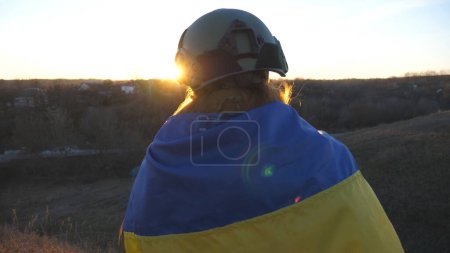 Unrecognizable ukrainian army soldier with flag of Ukraine looks at sunset. Girl in military uniform and helmet watch at horizon and wait for victory against russian aggression. Invasion resistance.