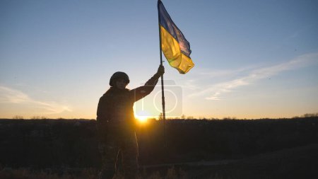 Frau in Militäruniform steht gegen Sonnenuntergang und hisst Flagge der Ukraine. Ukrainische Soldatin mit schwenkender Fahne. Sieg über die russische Aggression. Invasionswiderstandskonzept.